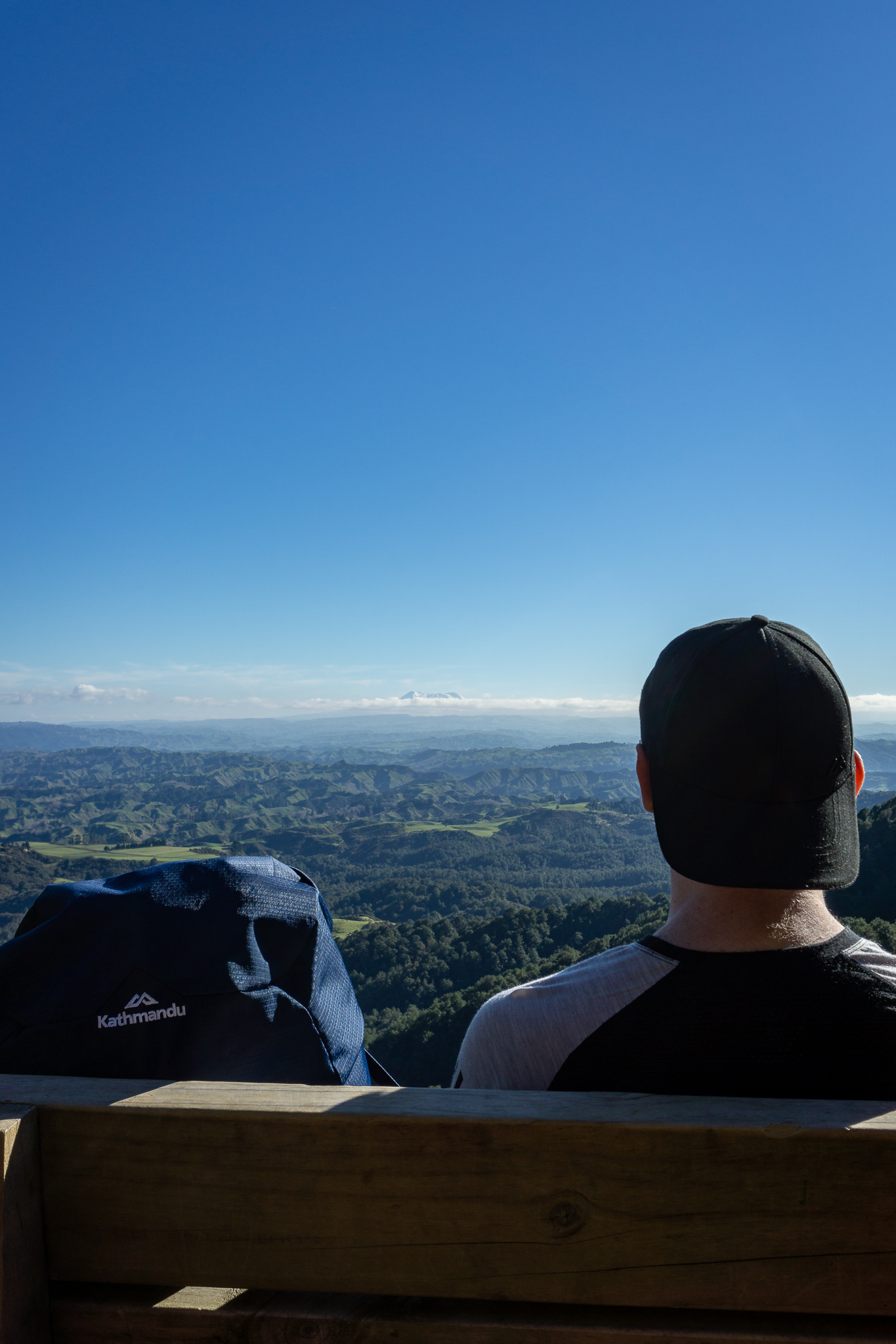 Close-up shot of a hiker wearing a cap sitting on a bench next to his Kathmandu pack with views over forest, rolling hills and Mt Ruapehu in the distance