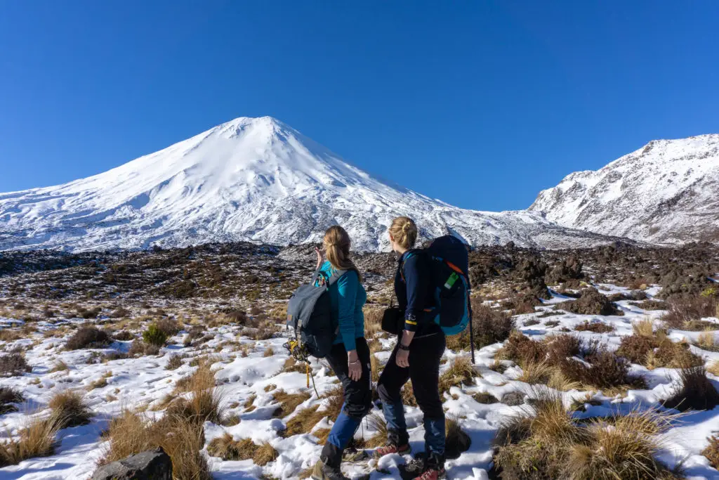 Two female hikers standing in snow-covered tussocks looking in awe at Ngauruhoe on the Tongariro Crossing in winter