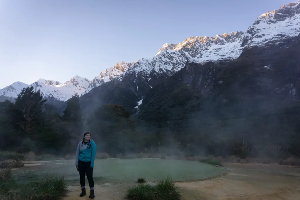 Female tramper standing next to the natural Welcome Flat hotpools at dawn with the morning sunlight hitting the tops of the snowcapped mountains