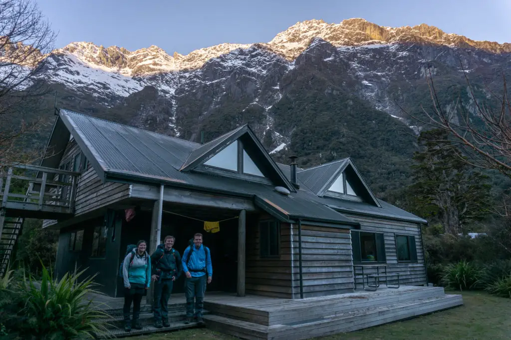 Three trampers standing on the deck of Welcome Flat Hut at dawn with the rising sunlight hitting the tops of the snowcapped mountains