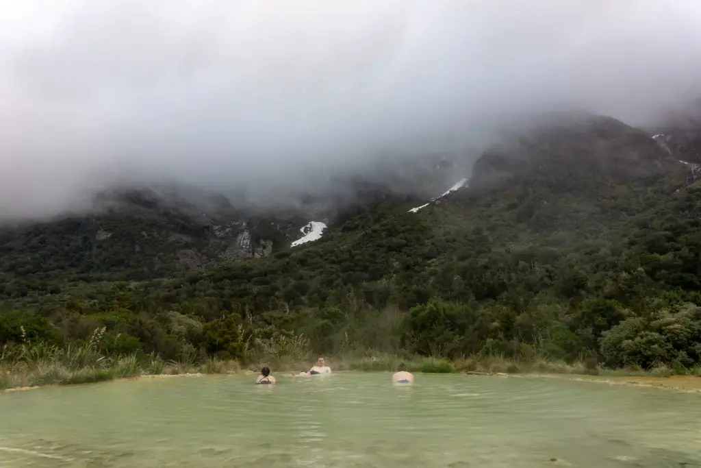 Three happy trampers soaking in the Welcome Flat natural hot pools teaming with low cloud covering the mountains in the background