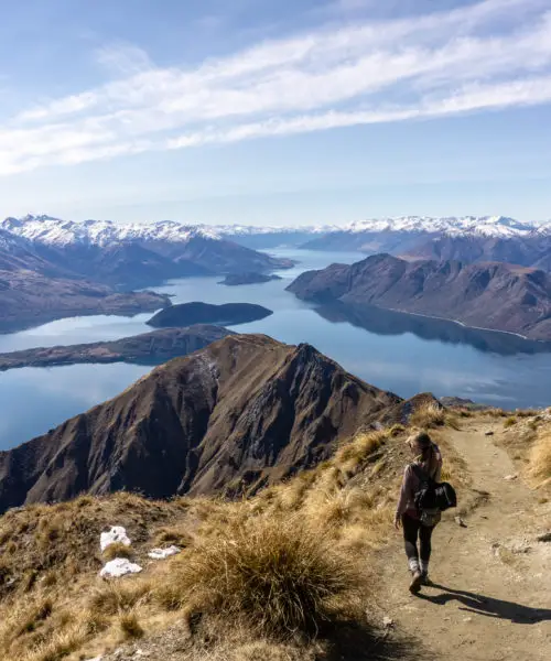 Female hiker walking down from the summit of Roys Peak track with Lake Wanaka, and mountains in the background