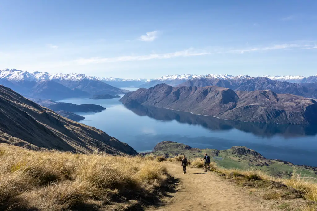 Two walkers hiking down Roys Peak track with a view of the surrounding mountains and Lake Wanaka in the background