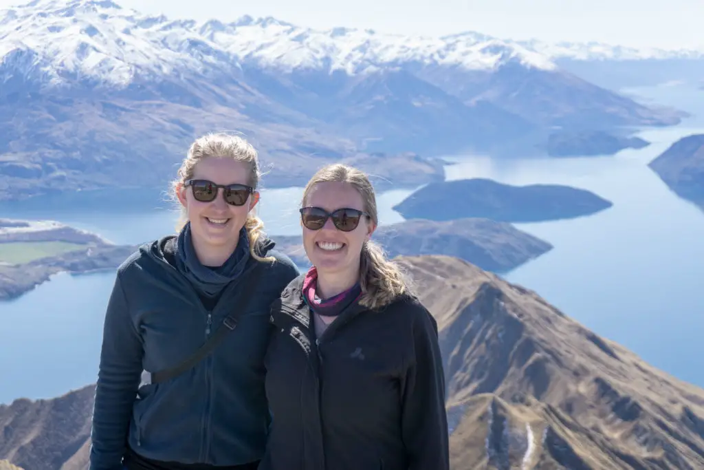 Two female hikers smiling while standing on the top of Roys Peak with Lake Wanaka and snowy mountains in the background