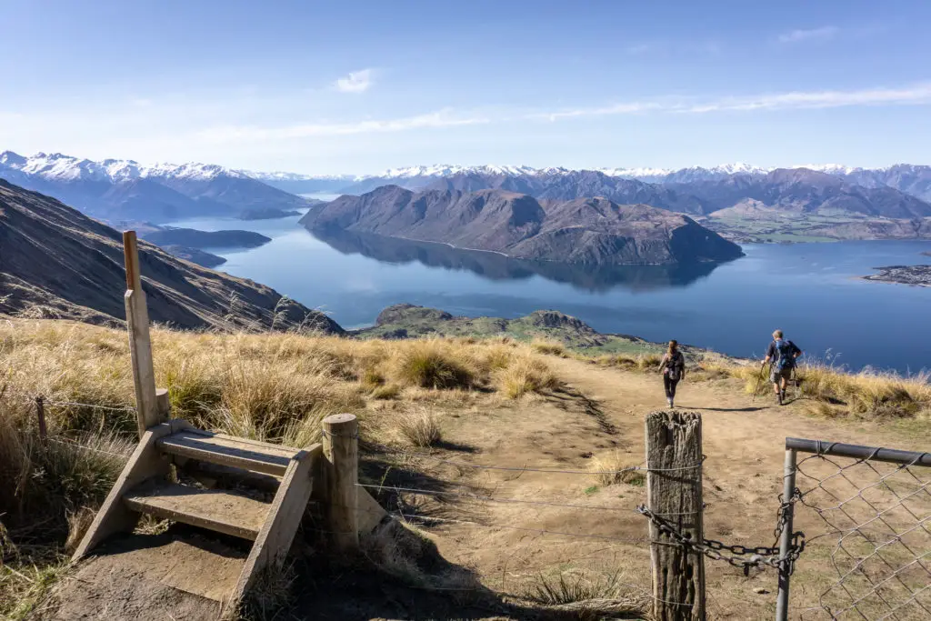 Style over top of a gate and fence on the Roys Peak track with two hikers walking down the trail and Lake Wanaka and mountains in the background