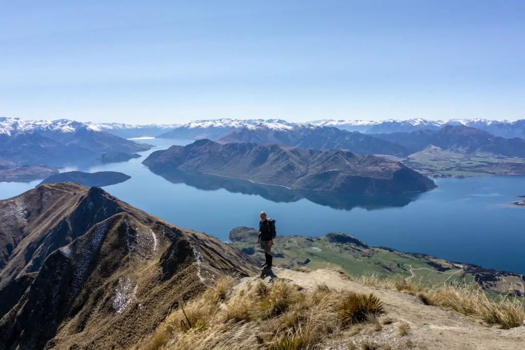 Male hiker standing on a viewpoint on Roy's Peak track with Lake Wanaka and snowy mountains in the background