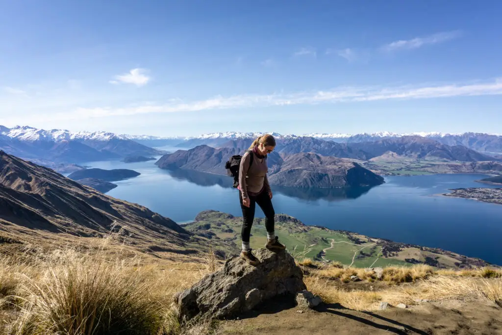 Female hiker standing on rock next to the Roys Peak track with Lake Wanaka and mountains in the background