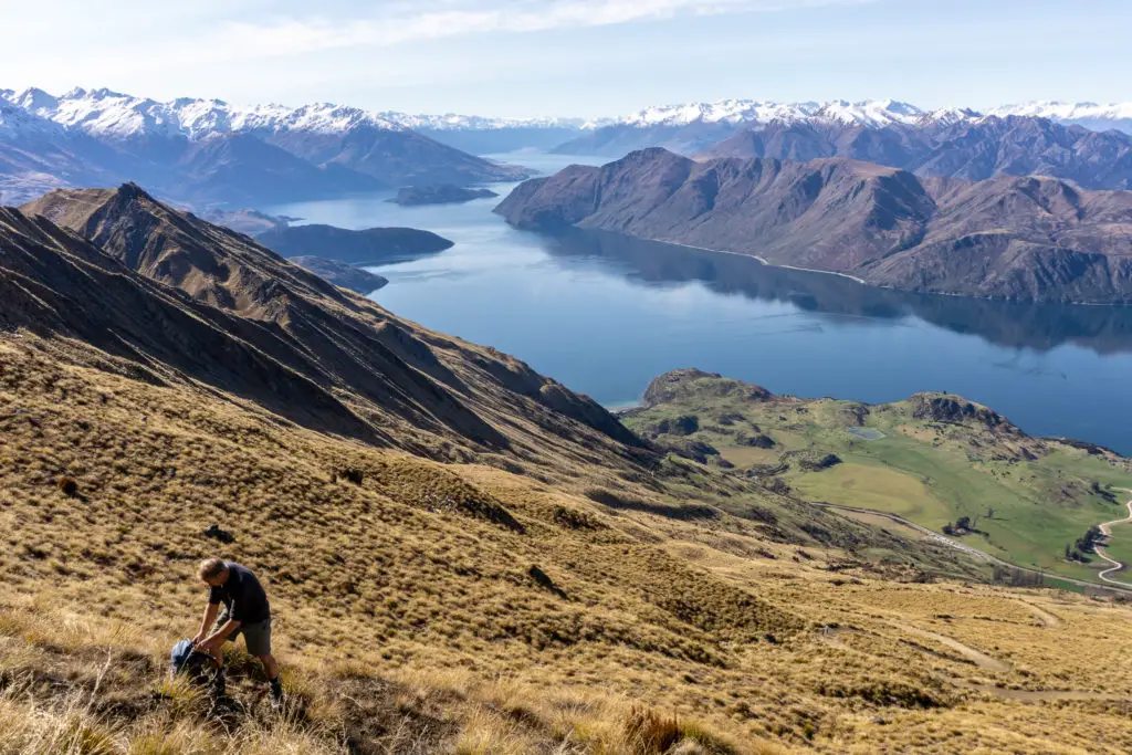 Male hiker fixing his pack on the tussock slopes of Roy's Peak with Lake Wanaka and mountains in the background