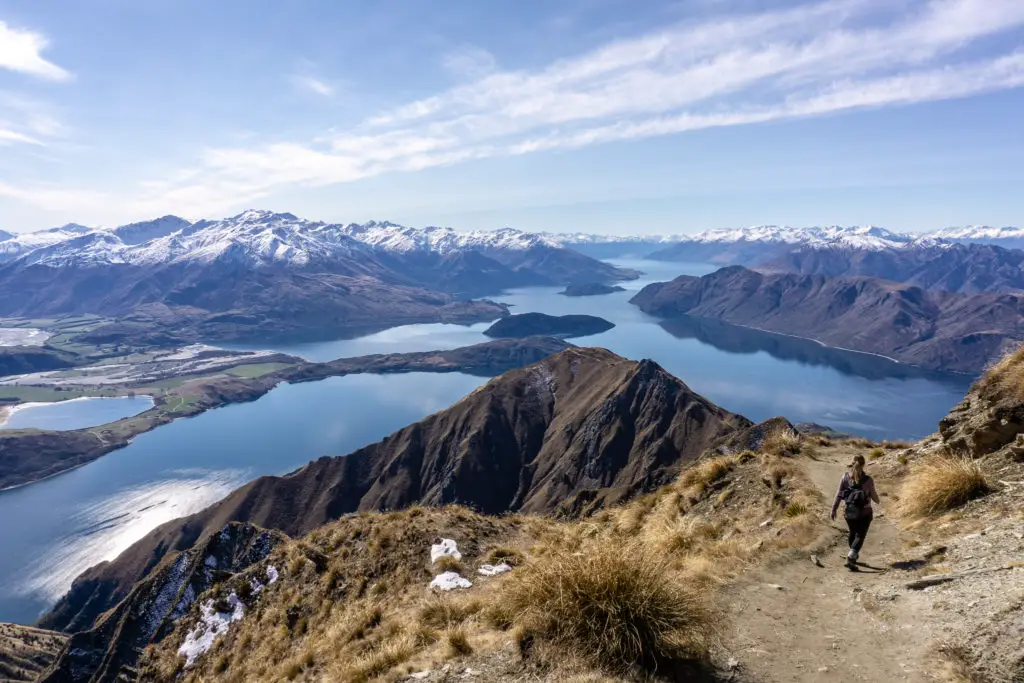 Female hiker walking down from the summit of Roys Peak track with Lake Wanaka, Glendhu Bay and mountains in the background