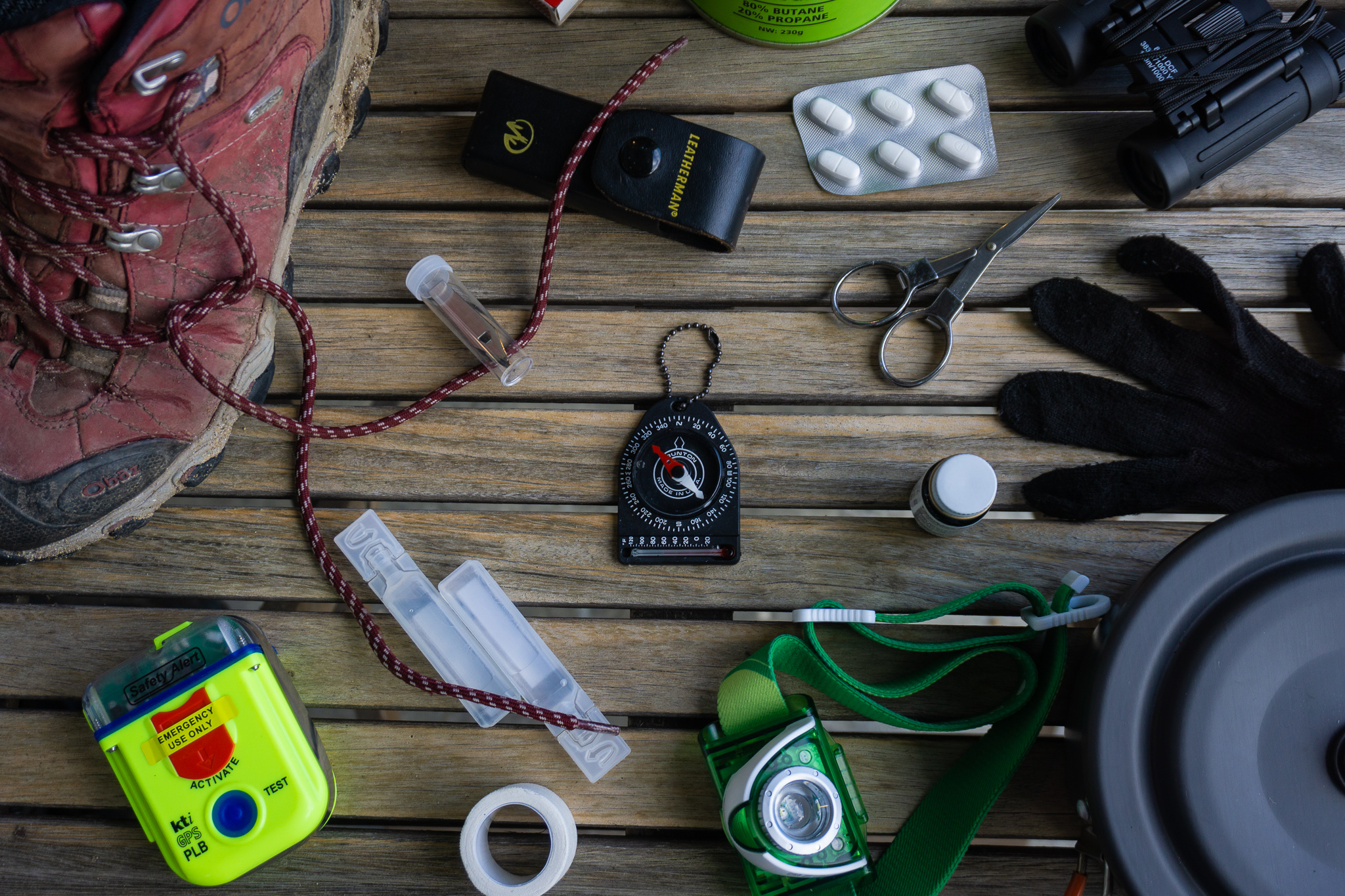 Flatlay of tramping / hiking gear with a compass in the middle, and boots, gloves, headlight, scissors, PLB, etc surrounding it