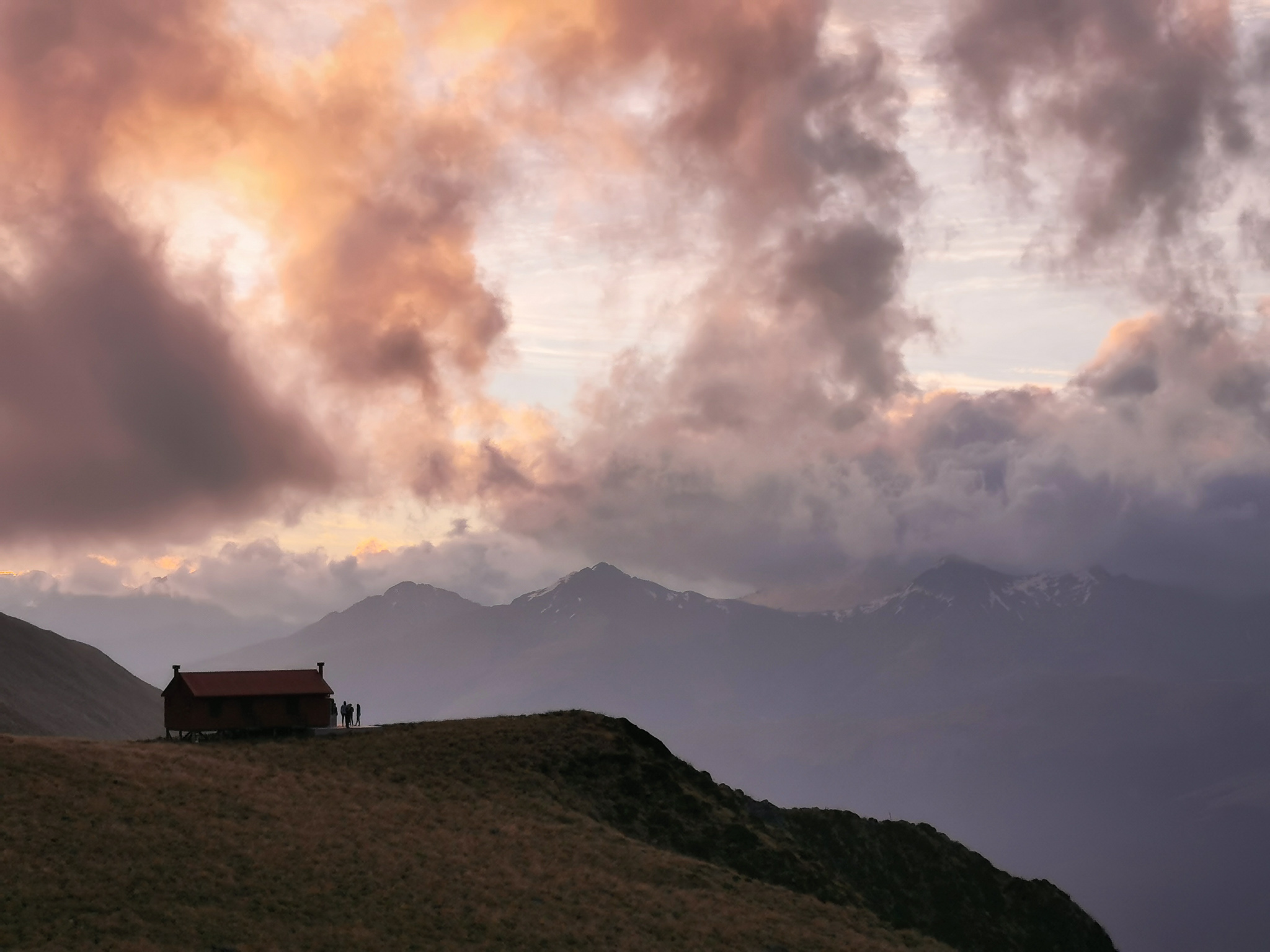 Brewster Hut with people standing on its deck silhouetted against a fiery sunset