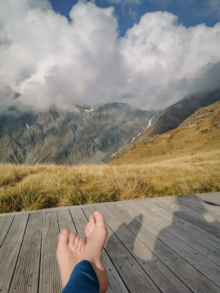 Bare feet sitting on the deck of Brewster Hut with tussocks, waterfall and mountains in the background