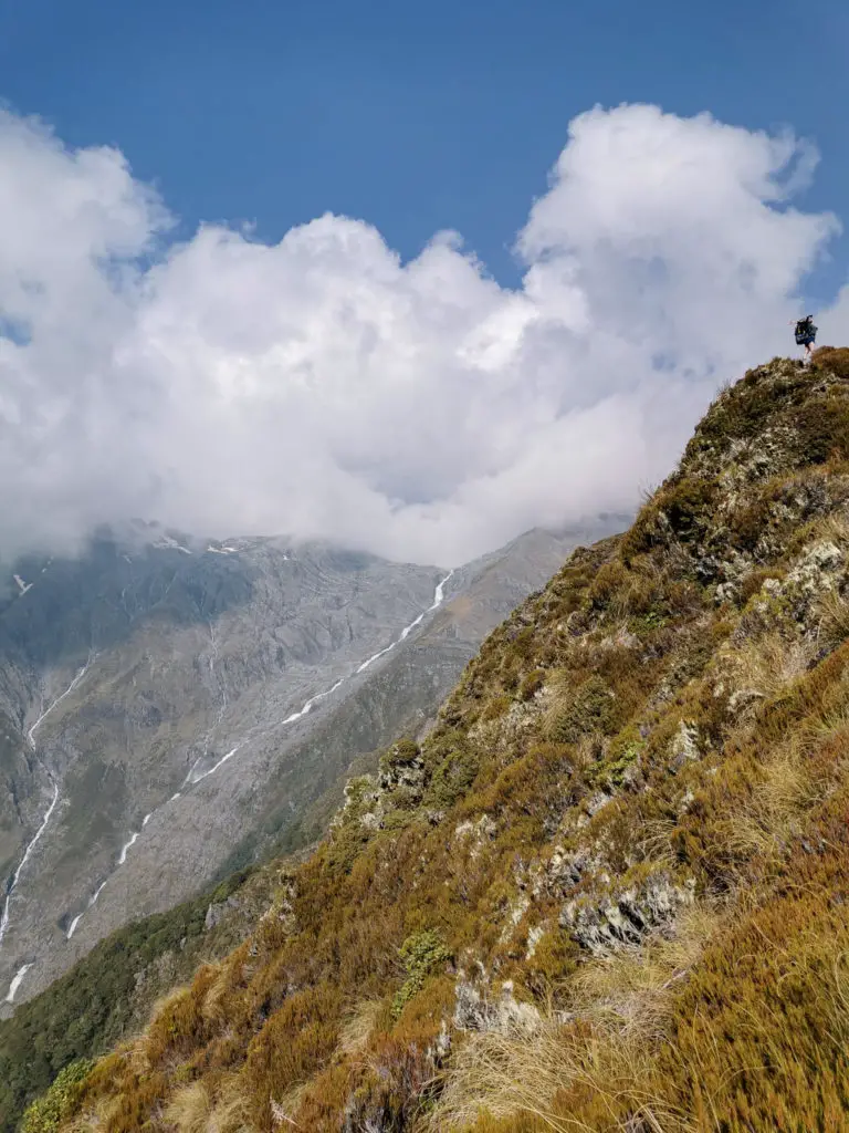 Hiker standing on top of a drop off on the way to Brewster Hut, waterfall and steep cliffs in the background