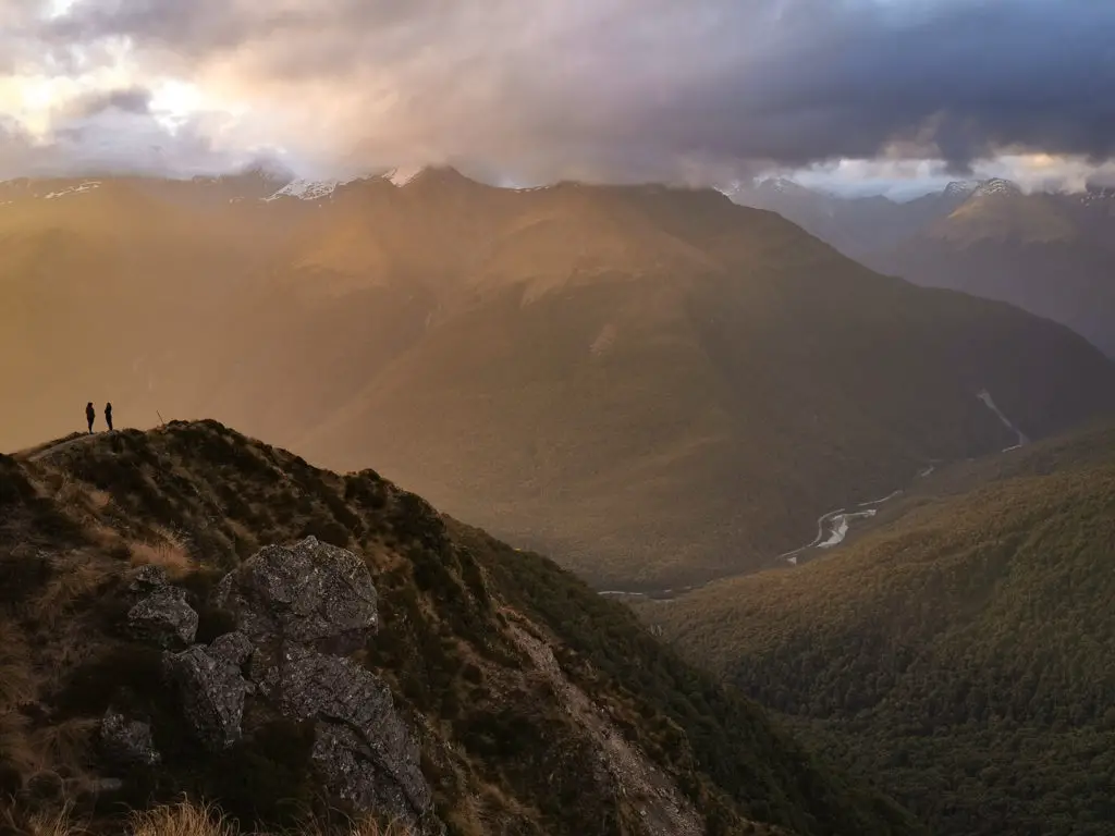 Two hikers silhouetted on the ridgeline with golden sunset light surrounding them and the Haast River winding through the valley far below