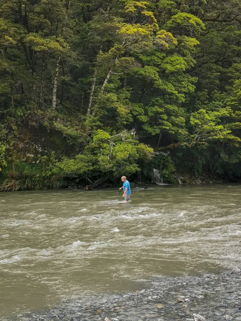 Man holding a pole crossing the flooded Haast River midway through with water above the knees