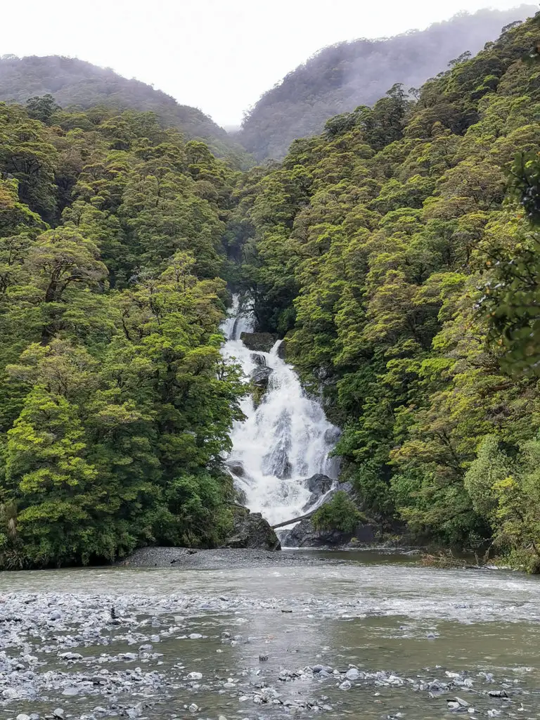 Fantail Falls cascading down in full flow with mossy trees on either side and the flooded Haast River in the foreground