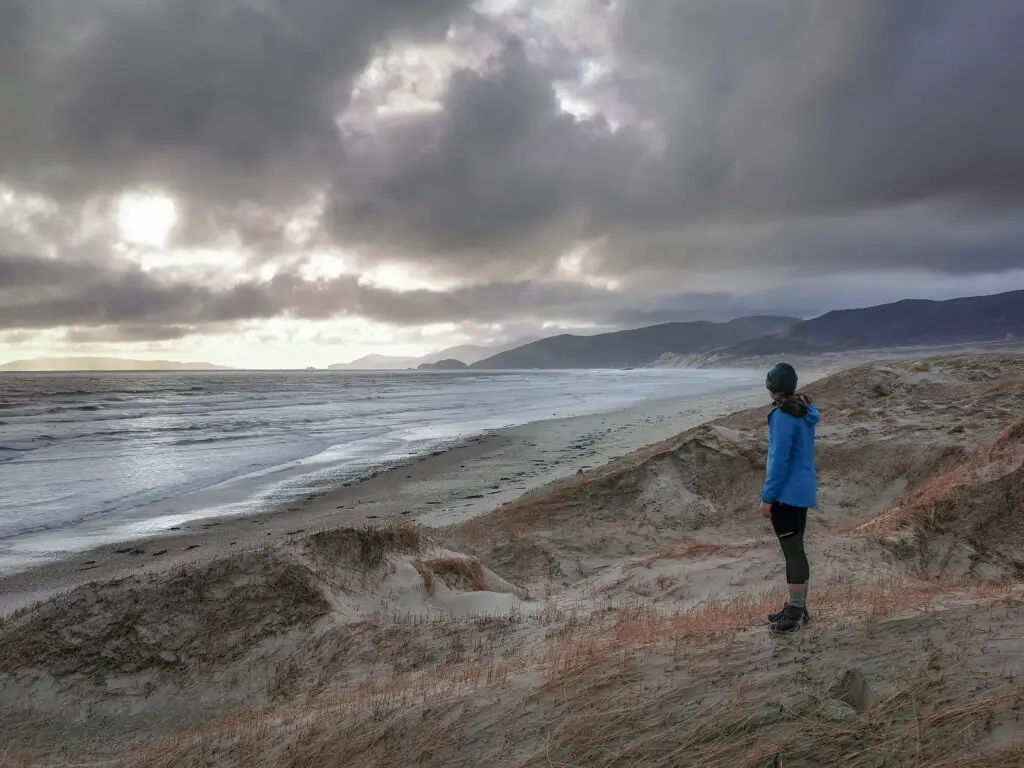 Hiker standing in the dunes overlooking Mason Bay looking up the coast, staying warm in winter