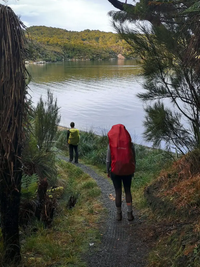 Two trampers with packs bearing colourful rain covers walking along a gravel track towards a bay