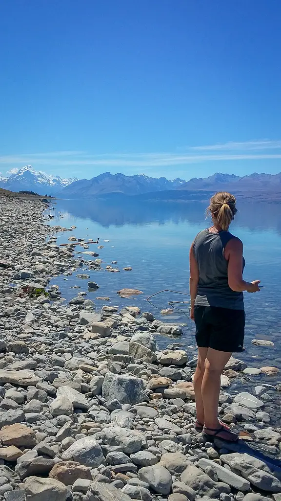 Lady on the edge of icy blue Lake Pukaki looking towards a distant Aoraki / Mt Cook