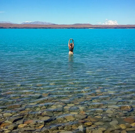 Lady standing in icy blue Lake Puakaki holding her arms up to the clear sky