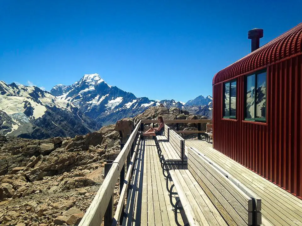 Lady sitting on a bench outside Mueller Hut reading a book with Aoraki / Mt Cook in the background