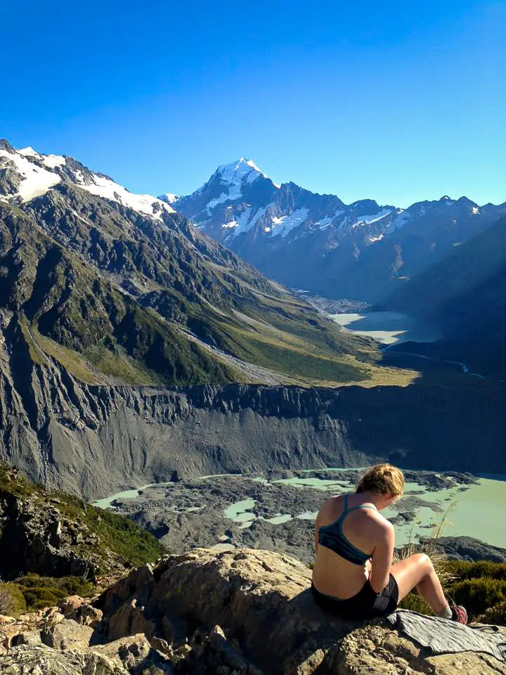 Lady wearing sports bra and shorts sitting on a rock with Mueller Lake, Hooker Lake, and Aoraki / Mt Cook in the background