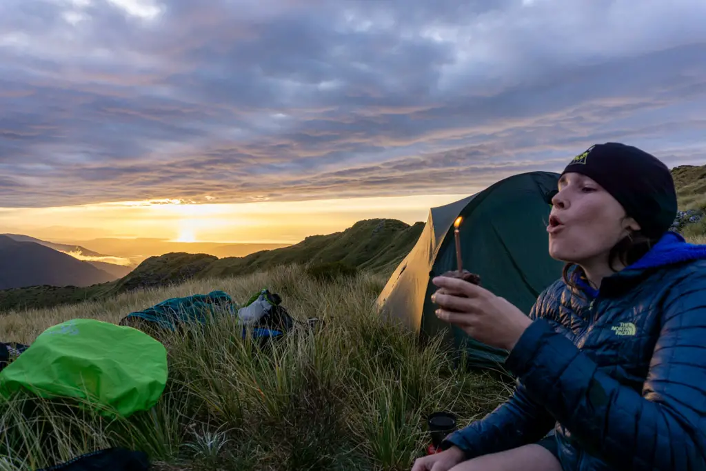 Hiker blowing out a birthday candle in front of a tent, while the sun sets over the Tasman Sea in the background