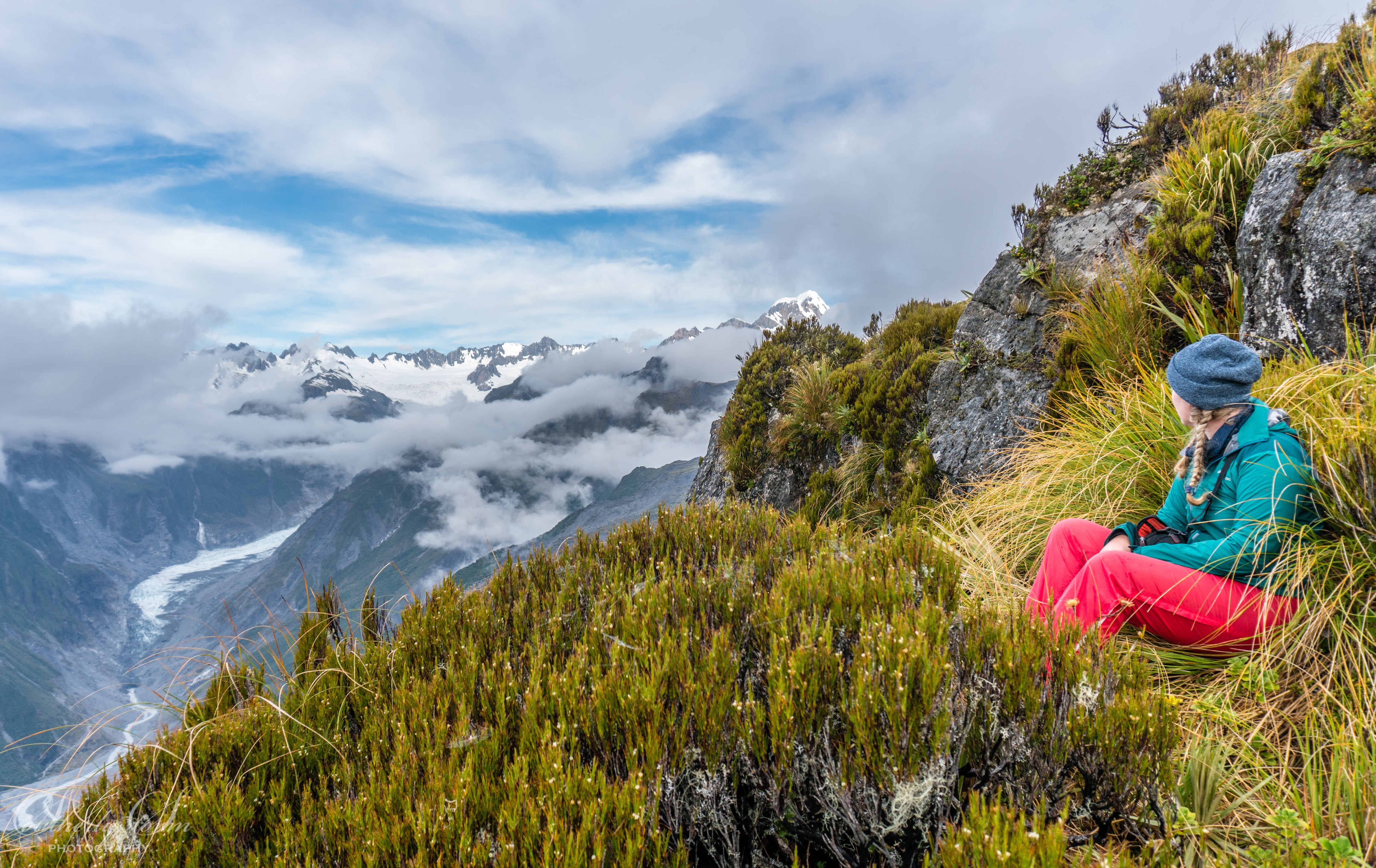 Woman hiker sitting in tussocks on Mount Fox looking out towards Fox Glacier
