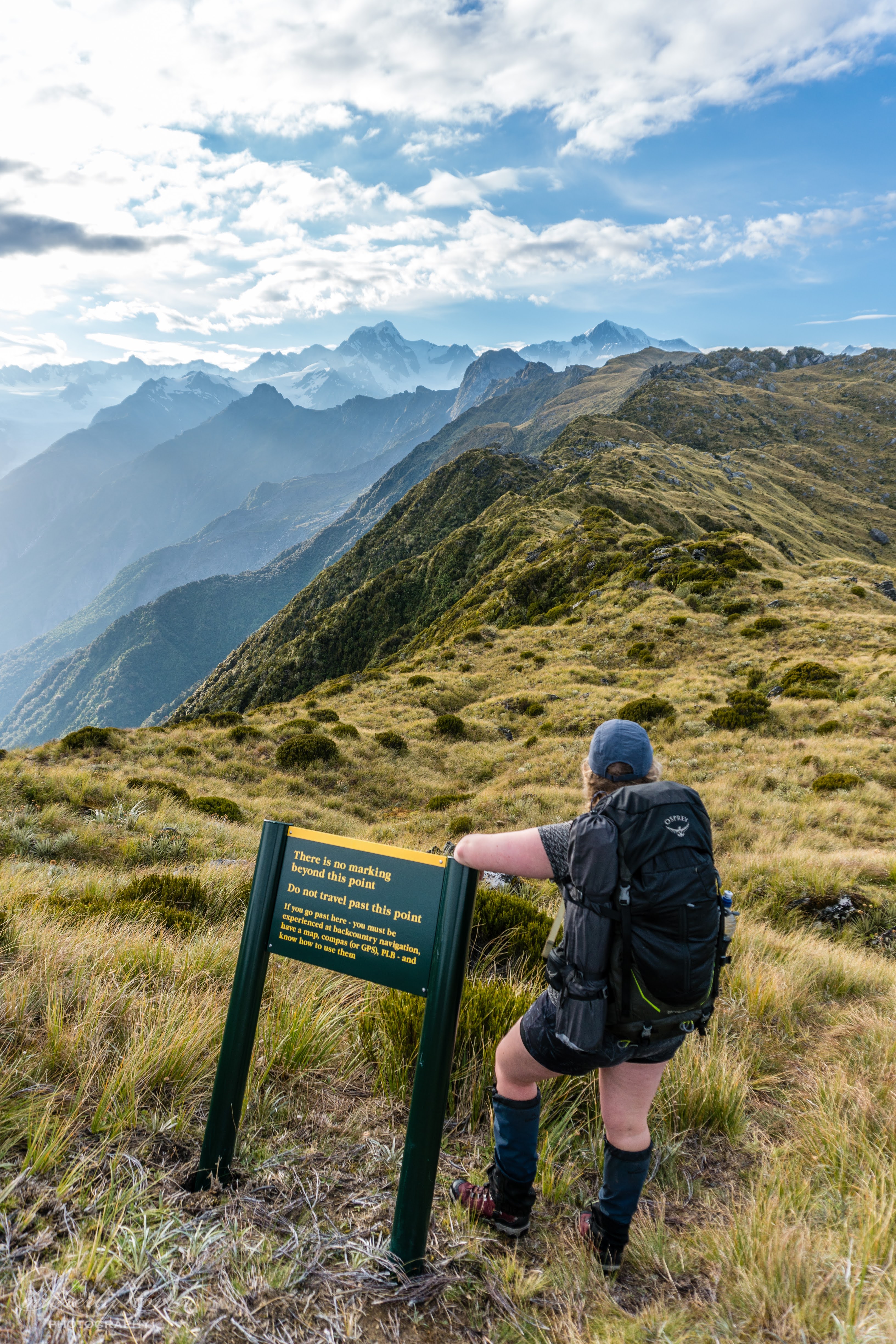 Female hiker standing by a DOC warning sign at the summit of Mount Fox overlooking Mount Tasman and Mount Cook
