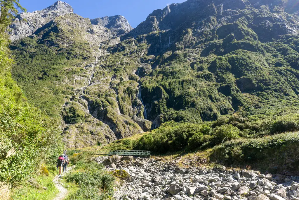 Hikers walking over a bridge on the Milford Track with towering cliffs and waterfalls above them