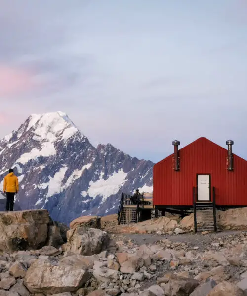 Mueller Hut with man in yellow jacket standing on a rock next to the hut and Aoraki in the background