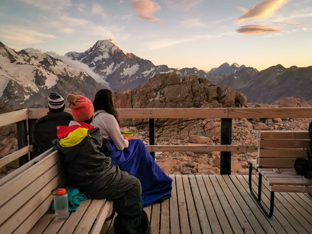 Adults sitting on the deck of a tramping hut in sleeping bags looking across to Aoraki Mt Cook at sunrise