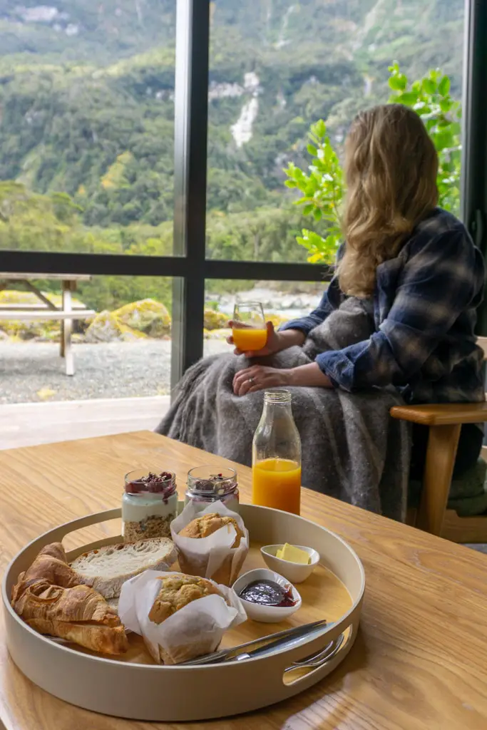 Woman sitting on chair in Milford Sound Lodge looking out the window with a delicious continental breakfast spread on the coffee table in front of her