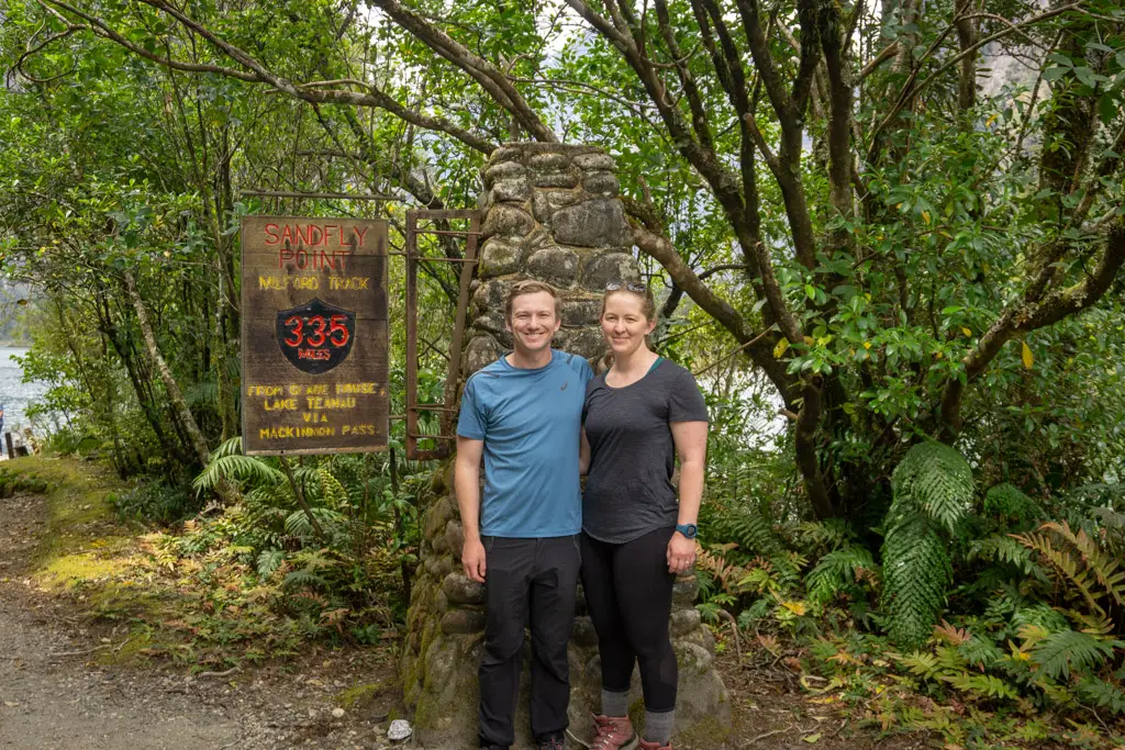 Two hikers standing in front of the sign as Sandfly Point marking the end of the Milford Track