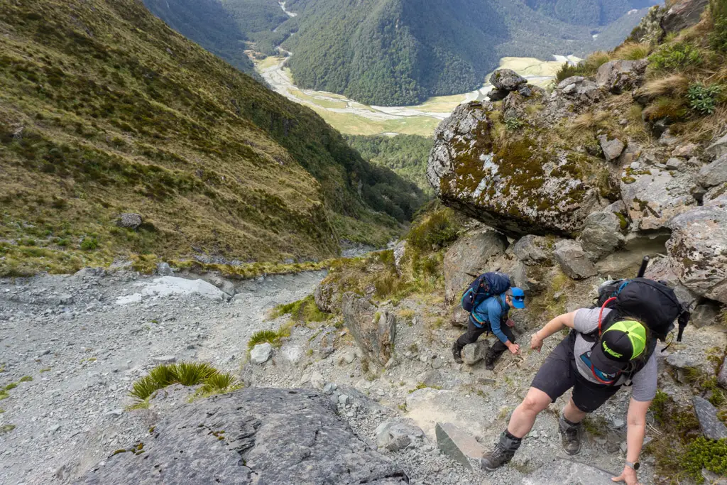 Two trampers climbing up a steep slip out of Emily Creek with Routeburn Flats far below in the background