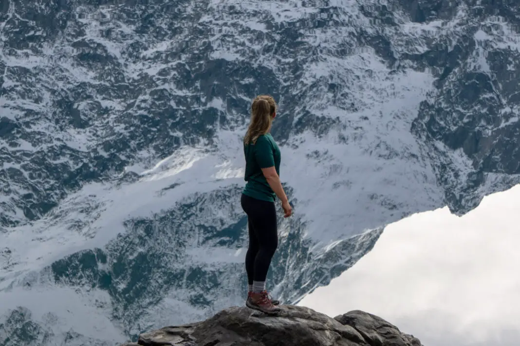 Woman standing on a rock in front of a lake reflecting snowy mountain peaks