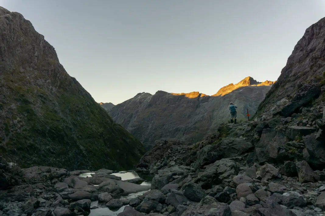 Man standing next to a small stream surrounded by mountains at dawn in Fiordland National Park