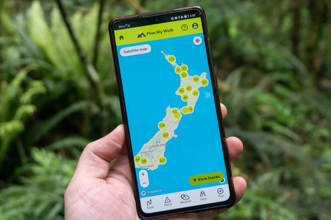 Hand holding a phone with Plan My Walk app open showing a map of New Zealand with various walks. Ferns and greenery in the background.