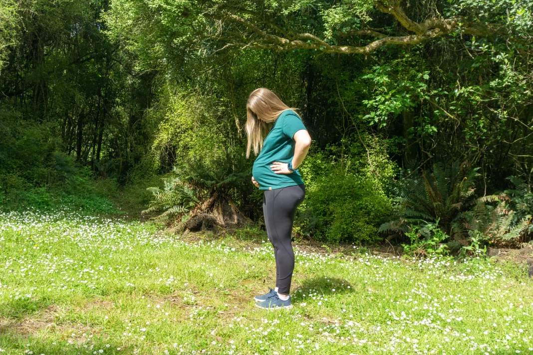 Pregnant woman standing in flowery field holding belly with trees behind