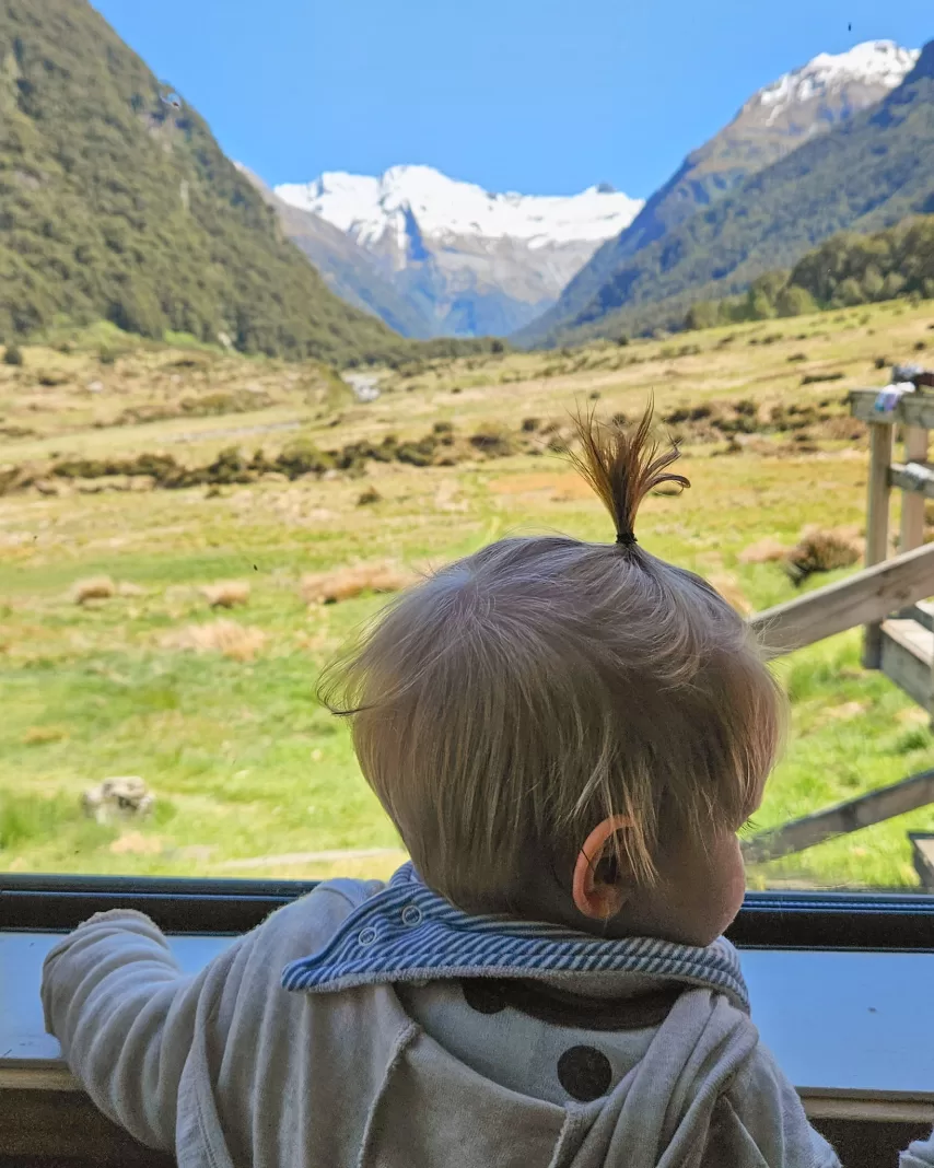Baby looking out the window of a tramping hut with snowy mountains in the background