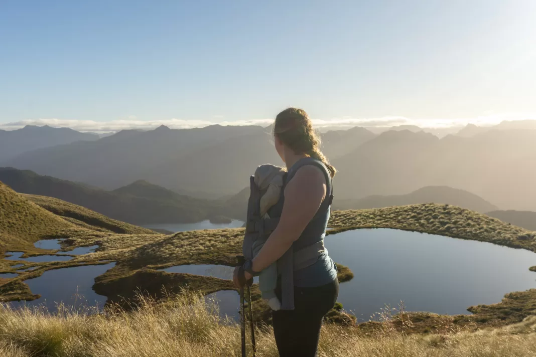 Woman carrying a baby on her front in a soft carrier with mountain tarns in the background at sunset