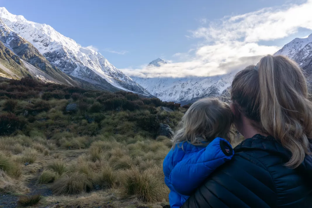 Woman holding her toddler with a Morrison Outdoors Co sleeping bag, with Aoraki Mt Cook in the background