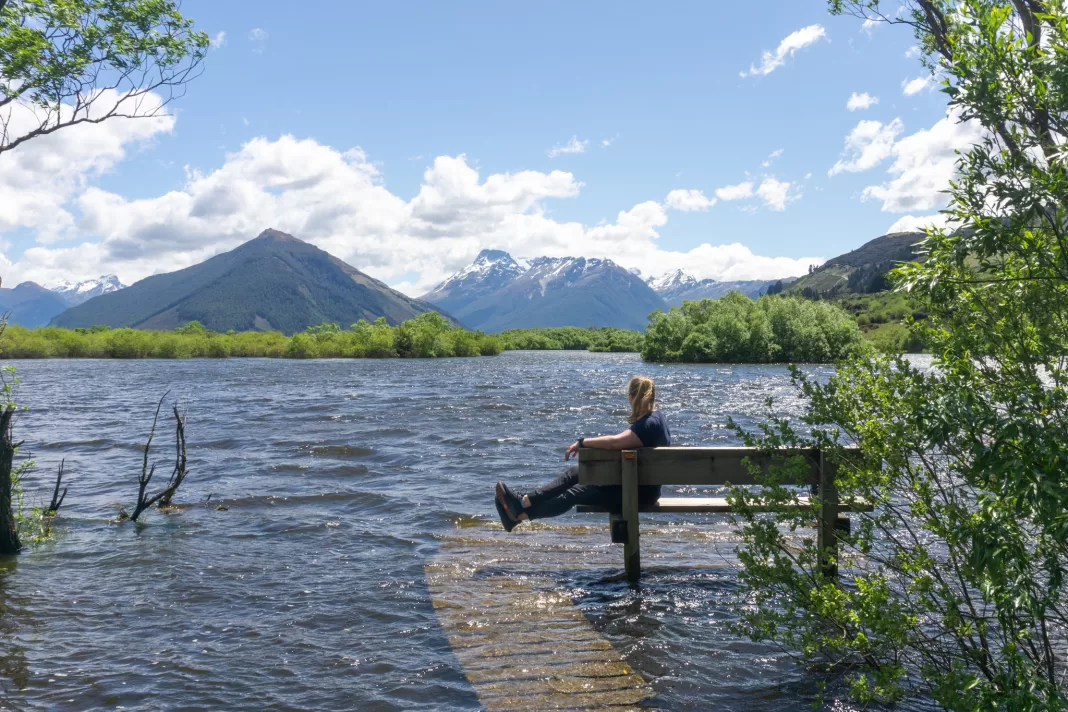 Woman sitting on seat on a flooded boardwalk with a lagoon, trees and mountains in the background