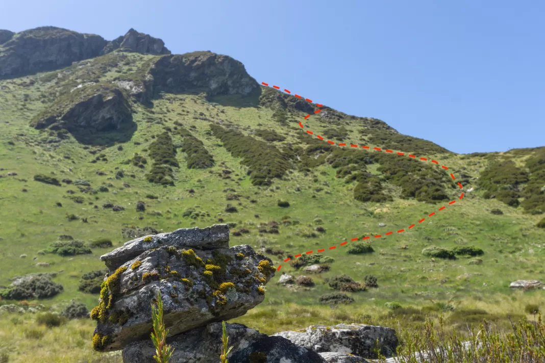 Photo of a cairn in the foreground, with a dashed red line showing the route up the grassy slopes that are out of focus behind it