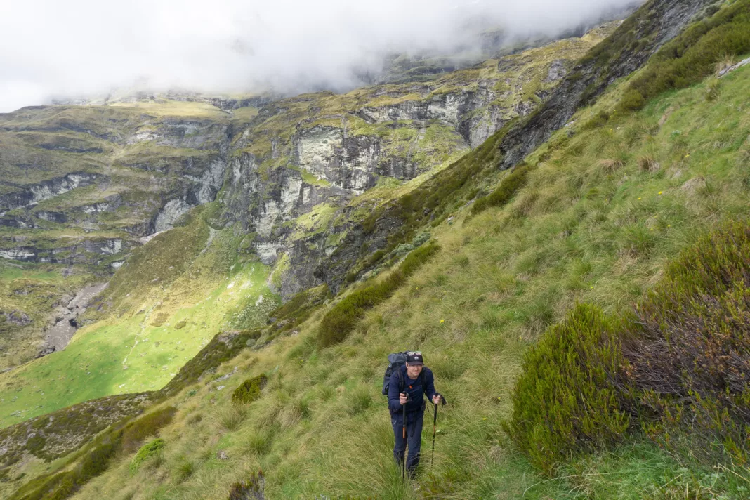 Tramper climbing up steep tussock slopes with cliffs and cloud behind him