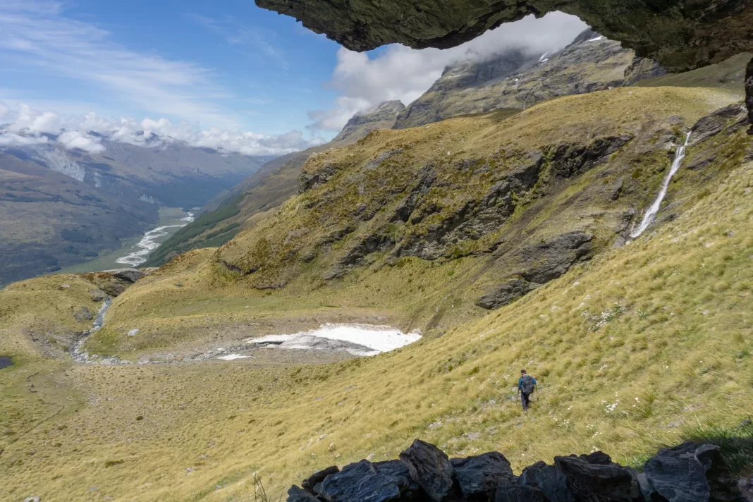 Photo taken from the upper bivy on the route to Mt Earnslaw, with a tramper walking down a tussock slopes with waterfalls, cliffs, mountains and snow in the background