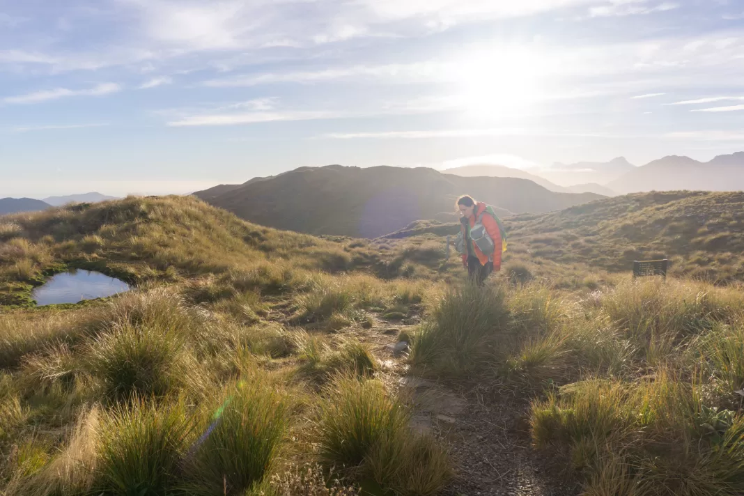 Woman walking through tussocks with a mountain tarn and peaks behind her at sunrise