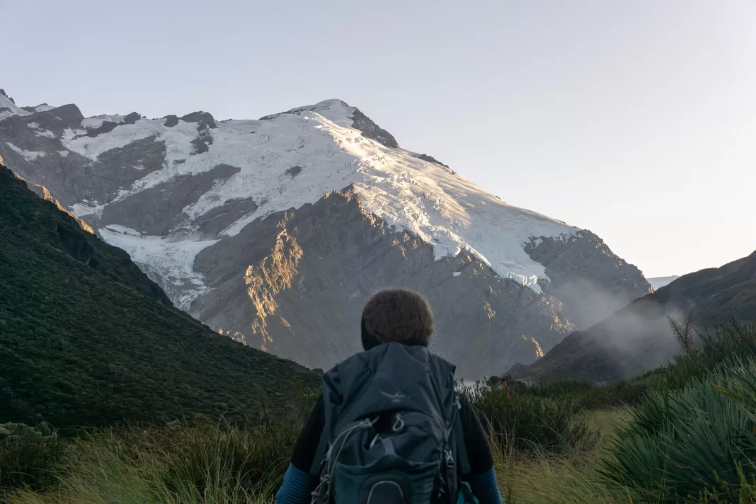 Woman with a pack on her back looking towards snowy mountains lit by sunrise