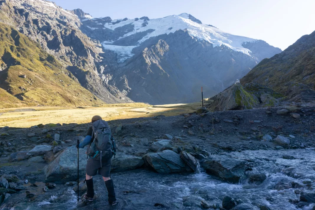 Woman crossing a stream with Mt Edward and the Marshall and Hesse Glaciers in the background