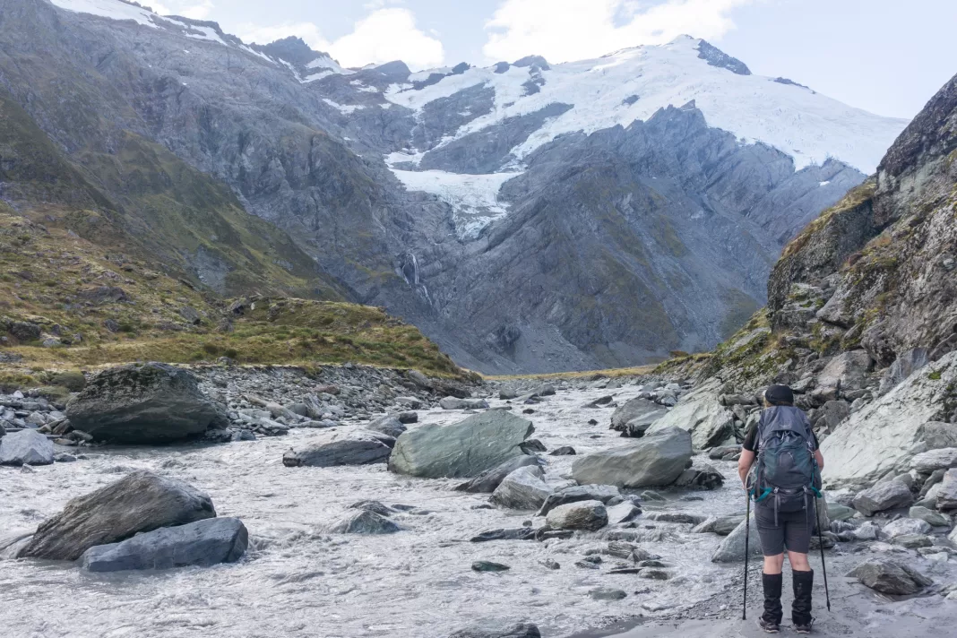 Tramper standing next to a milky river looking at mountains and glaciers in the background